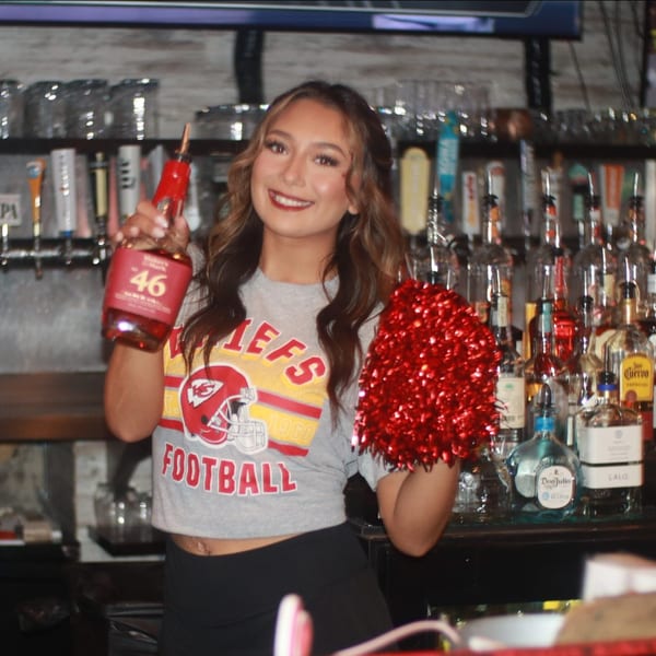 Smiling bartender in a Chiefs shirt holds a bottle of Maker's Mark and a red pom-pom.