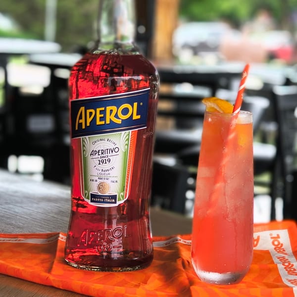 A bottle of Aperol next to a tall glass of Aperol Spritz with an orange slice and straw.
