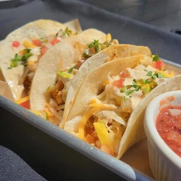 Three American-style tacos with cheese, salsa, and sauce, served with a side of salsa.